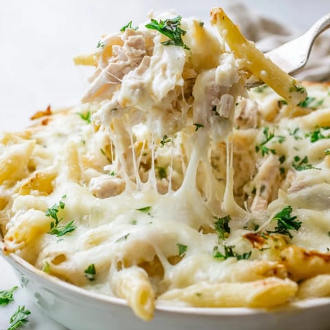 Fork lifting creamy Chicken Alfredo Bake with tender chicken and penne pasta, steam rising.